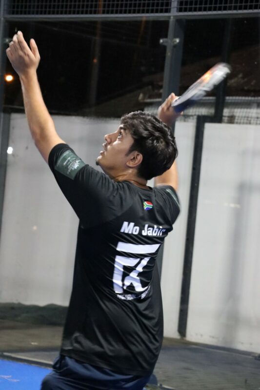 A man is playing paddle. Both of his arms are in the air; it looks like he is about to hit the ball. He is holding his racquet in his right hand. His back is toward the camera and you can see white writing on his black shirt; as well as the South-African flag. There is a white wall behind him. He is playing at night.