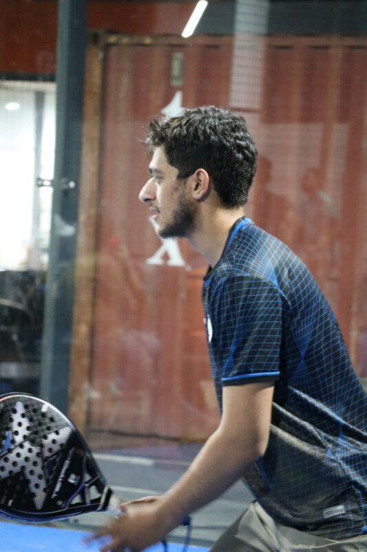 A man is playing paddle. The photo is taken from the side and the man is wearing a blue shirt. He has dark hair and a beard. There is a brown container behind him. He is playing at night.