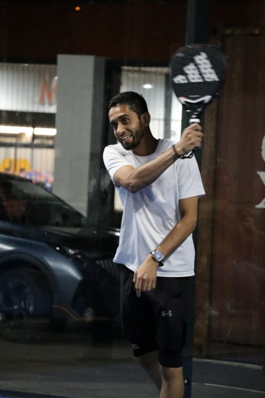 A man is playing paddle. His racquet is close to his head. He is holding it with his right hand. He is smiling and looking in front of him. The man is wearing a white shirt and black pants. There is a car in the background. He is playing at night.