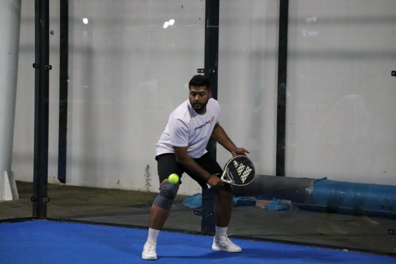 A man playing paddle. He is about to hit the ball. He is wearing a white shirt with black pants and white socks and shoes. The court surface is blue. He is playing at night time. There is a white wall behind him. His legs are bent.