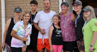 Members from the Newcastle squash club posing for a photo after their sucessful completion of the 4 kilometer walk for cancer awareness. They are all wearing different coloured sporting outfits and holding their medals. The are standing in front of a facebrick building.