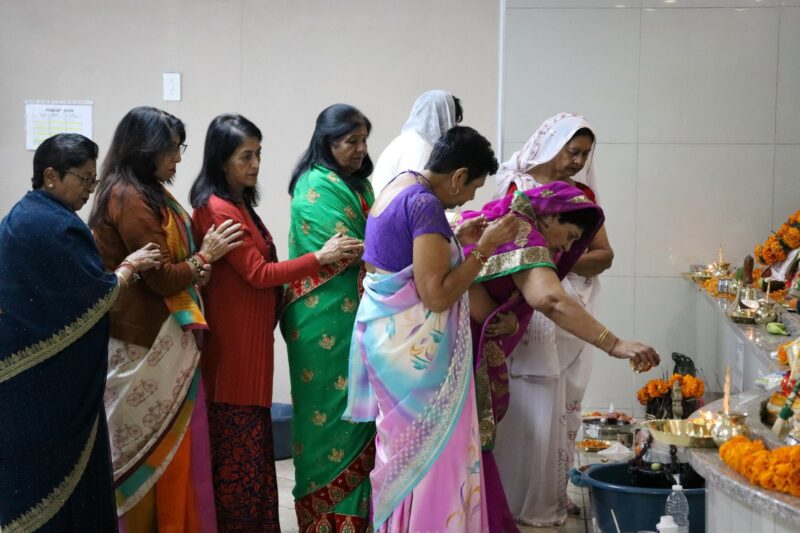 Devotees remained awake through the night, offering milk, sour milk, water, ghee and bilva leaves to the Shivalinga. These devotees are wearing multicoloured sari's and some are covering their heads. They are inside the temple. They are standing, facing the Shivalinga. The temple is white inside.