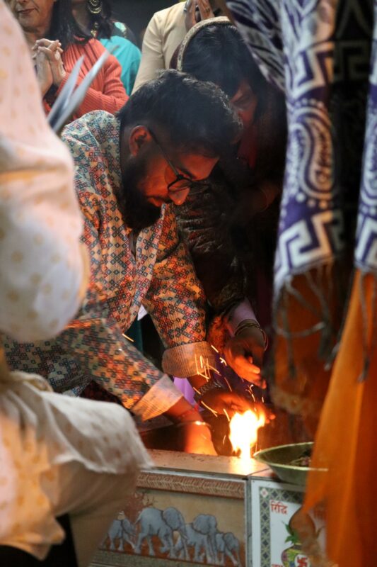 A devotee in a colourful shirt is making a fire. His hands are closely in the pit, surrounded by other people. He is wearing glasses and has dark hair and a long beard. It is night time.