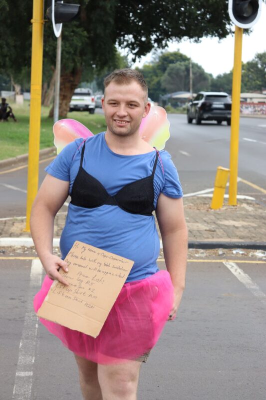A man wearing a blue T-shirt, black bra, pink tutu and fairy wings stands in the middle of a road holding a handwritten cardboard sign asking for donations.