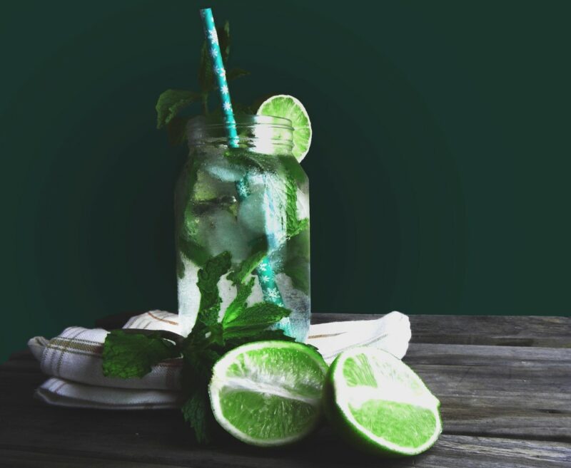 A green glass jar filled with liquid, limes and mint leaves on a dark table setting.