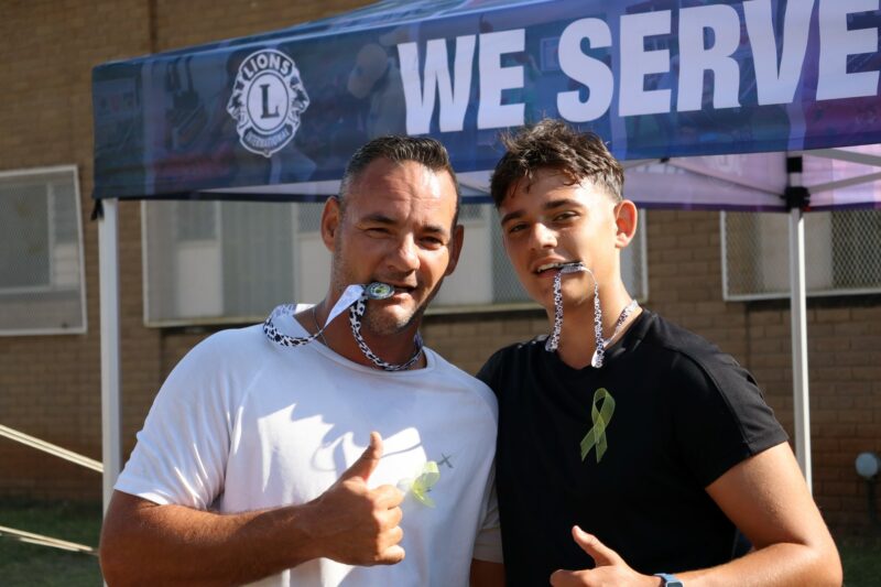 Father and son posing for a photo with medals in their mouths. They finished fourth and fifth in the Lions Club Big Walk for Little warriors cancer awareness walk. There is a gazebo in the background. It is a sunny day. They have yellow ribbons pinned to their shirts.