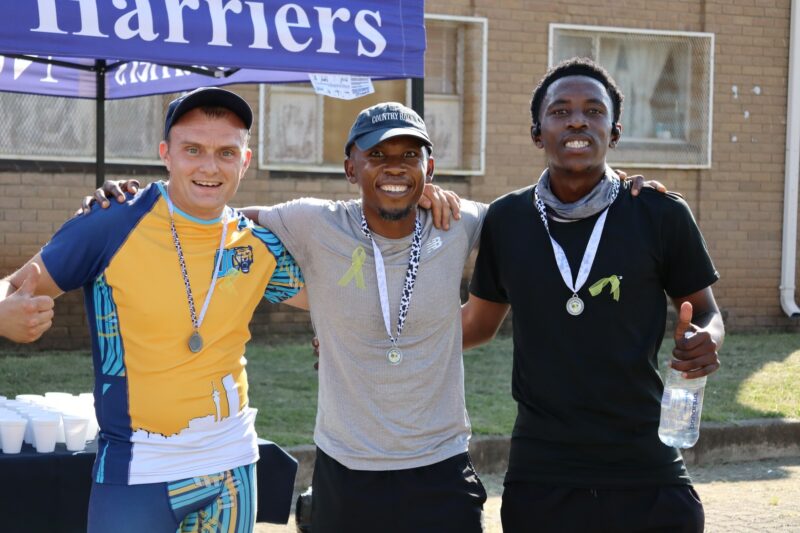 Three athletes smiling at the camera as they finished in the top 3 for the Lions Club Big Walk for Little Warriors.They have medals around their necks. It is a sunny day. The athletes are wearing athletic wear. There is a gazebo in the background.