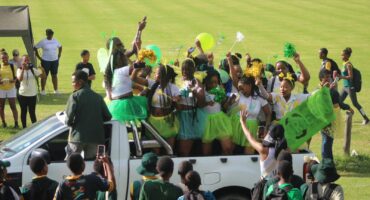 Image of learners dressed in cheerful white, green and yellow clothes while standing on the back of a white bakkie.