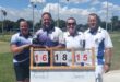 Four bowlers stand behind the score board posing for a photo in their club attire.