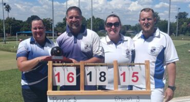 Four bowlers stand behind the score board posing for a photo in their club attire.