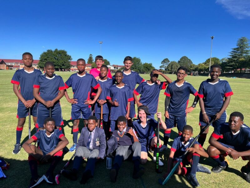 A group of school boys are posing for a photo. They are wearing sport attire for playing hockey. They have hockey sticks in their hands. It is very sunny outside and they are standing on a hockey field that is made of grass.