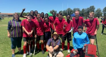 Volksrust High School boy's hockey team is posing for a photo. They are wearing red shirts; the goalie is wearing a blue bib. The boys are all smiling in the sun in Vryheid, where there hockey match took place. They are standing on the grass, with a teacher/coach sitting on the grass in front of them.