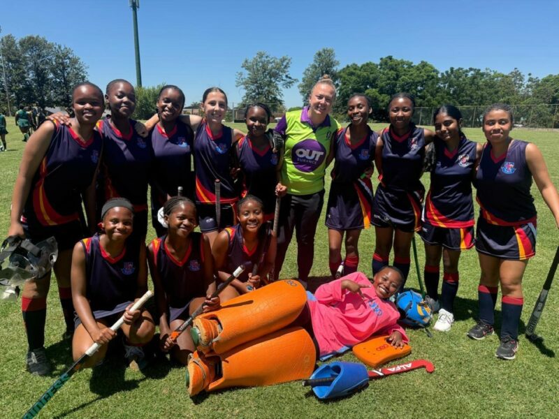 A group of Volksrust High School students in their hockey kits posing for a photo in Vryheid. The goalie is laying on the floor and she has oragne padding and a bright pink bib on. There is a coach/umpire in an Out Surance shirt that is white, purple and green. The students are holding hockey sticks and smiling. It is a sunny day with green grass.