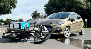 A vehicle and a deliver motorbike collided at an intersection in town. The vehicle is bronze and the motorbike is white, black and turqoise. Is is morning. There is fluid on the road.