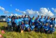 A group of anglers dressed in blue fishing attire pose for a photo outside in the sun. blue skies and green landscapes are pictured in the background.