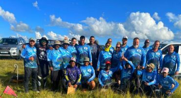 A group of anglers dressed in blue fishing attire pose for a photo outside in the sun. blue skies and green landscapes are pictured in the background.