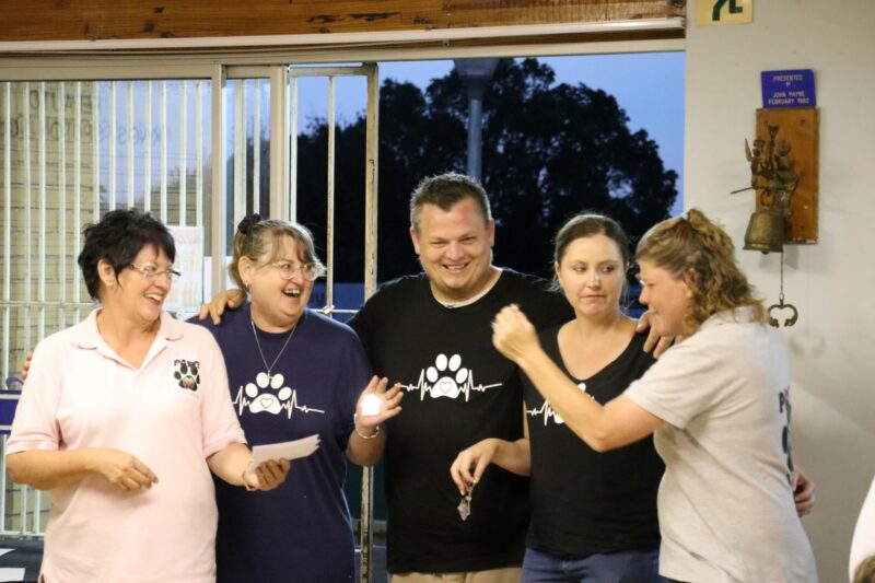 Alet Oberholzer handing a prize over to the winning team of the bowls competition. There are four members in the team. They are wearing shirts with animal paw printing on it. The people are inside a building, and it is dark outside.