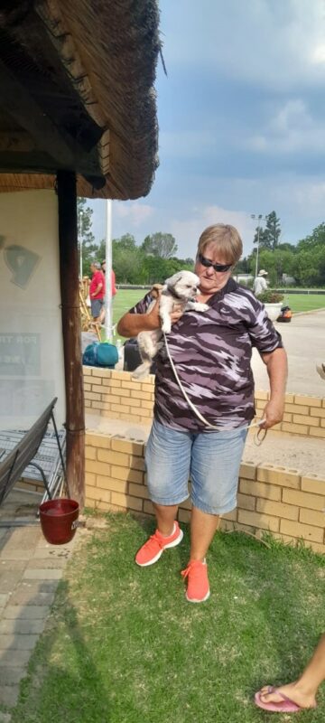 A woman wearing a busy shirt, is holding a white-ish dog under her right arm. She is looking down at the dog. The woman in standing in front of a small facebrick wall.