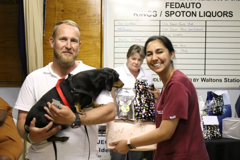 A man with his black daschund, next to the local veterinariam who is wearing maroon scrubs. There is another woman in the background, in from of a white board. The veterinarian is holding packages in her arms.