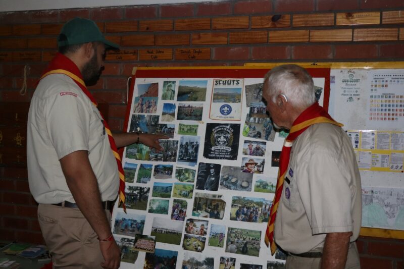 Two Scouts leaders in Scouts uniform standing in front of a board filled with photos of past camps and Scouting events. The one leader is pointing at a photo.
