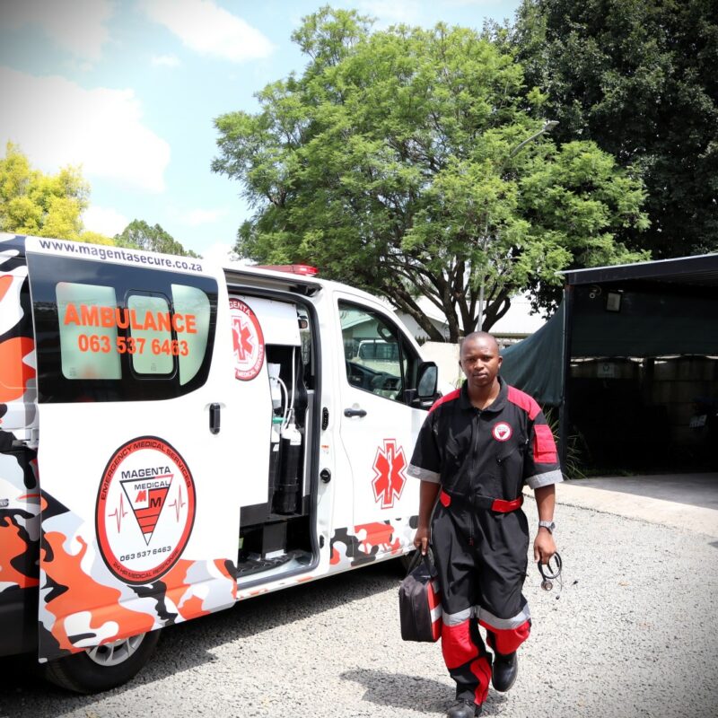 Sakhile is walking toward the camera. He is walking next to an ambulance that is white, black, and red; it has Magenta's logo on the door. He is wearing his paramedic uniform and holding a medical supply bag and stetascope in both hands. It is a sunny day outside.
