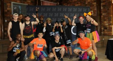 The top 3 teams at the Kyotech 2026 Lekker Resies. They are all posing infront of a Kyotech banner, all dressed in different, silly outfits. The team who came first is wearing orange with colourful tutus.