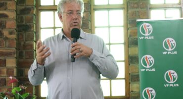 A man is standing in a building with facebrick walls in the background. There is a green banner with the word VF Plus printed on it. The man is speaking to a crowd, and he is holding a microphone in his left hand. His right hand is in the air.