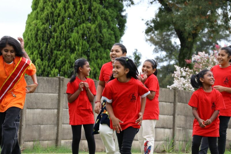 Indian children running outside. They are all wearing red t-shirts. The grass is green. There is a concrete wall in the background.