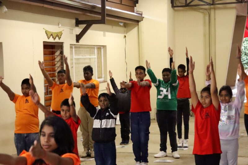 A group of children with their hands in the air. The children are standing in a straight line. There is a beige wall in the background. The children are indoors.
