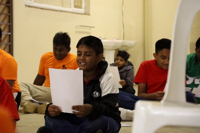 A boy is holding a piece of paper in front of him. He is smiling while singing a traditional Hindu song. All the children in the background are doing the same thing. They are indoors.