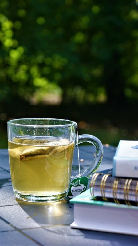 A cup of chamomile tea with a tea bag inside. The tea cup is next to some books on a table outside. There is a string attached to the tea bag. The cup, books, and table are outside.