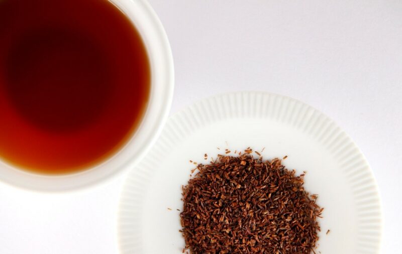 A cup of Rooibos tea in a white polistirene cup. There are Rooibos leaves next to it, in a white paper place.