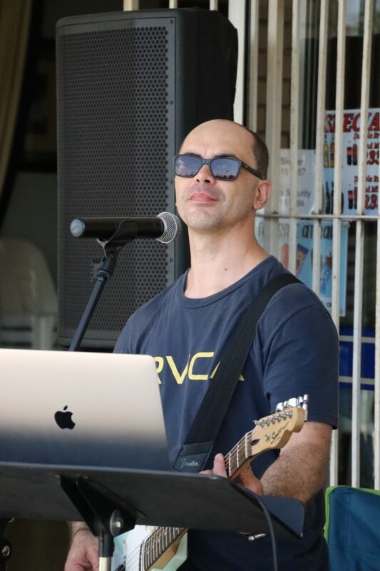 Singer and musician, Jaco Strydom behind the microphone and his laptop; singing and playing the electric guitar. He is wearing a blue t-shirt and sunglasses.
