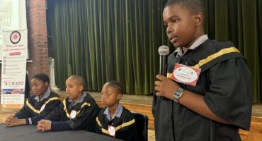 Young learners dressed as magistrates/judges sitting behind a table with one learner holding a microphone.