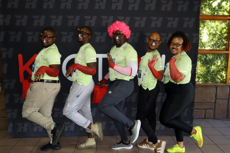 A Lekker Resies team dressed in green shirts with animal snouts attached to their fake glasses. The tall man in the middle is wearing a pink clown wig. All of them have red sleeves on their arms, with their left legs bent.