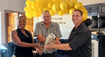 Image of a woman and two men, with yellow balloons in the background while the one man and the woman hand gifts to the second man.