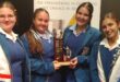 Image of learners in blue uniforms and white sashes holding a trophy.