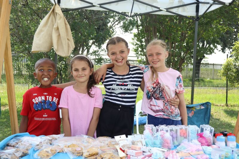 Four children smiling behind their stall at the Newcastle Bowling Club. They are standing under a gazebo with trees in the background.