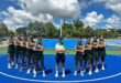 Image of first team netball players standing in a halfmoon with the coach on a blue court.