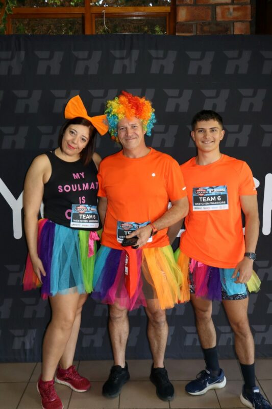 A family of three is standing in front of a big banner. The father and son is wearing orange, with the mother wearing an organce bow on her head. All three are wearing multi-coloured tutus.
