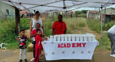 Grown-ups and children are photographed standing next to a table, while handing out food parcels in Siyahlala.