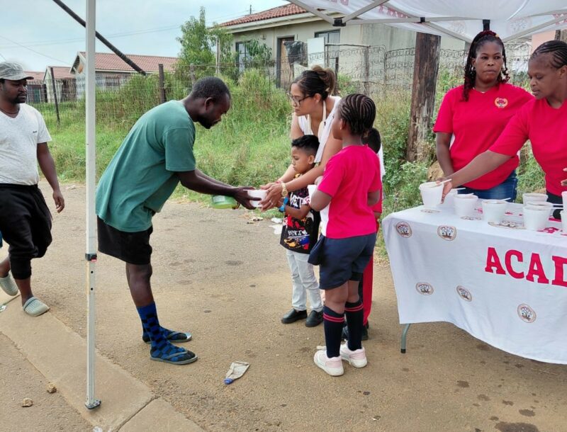 Grown-ups and children are photographed standing next to a table, while handing out food parcels in Siyahlala.