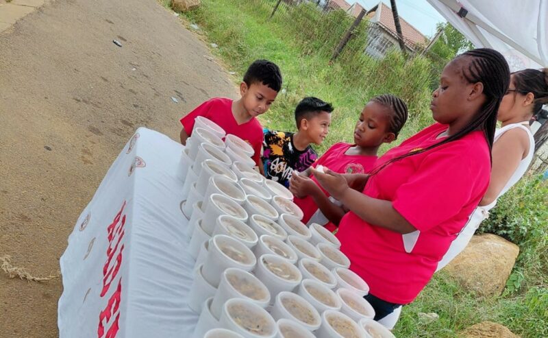 Grown-ups and children are photographed standing next to a table, while handing out food parcels in Siyahlala.