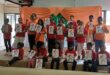 Children holding certificates at a local camp in Newcastle. They are wearing red and orange, with dark pants. The background is draped with orange and green cloth. Some children are sitting, while others are standing behind them.