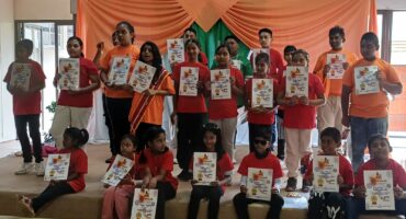 Children holding certificates at a local camp in Newcastle. They are wearing red and orange, with dark pants. The background is draped with orange and green cloth. Some children are sitting, while others are standing behind them.