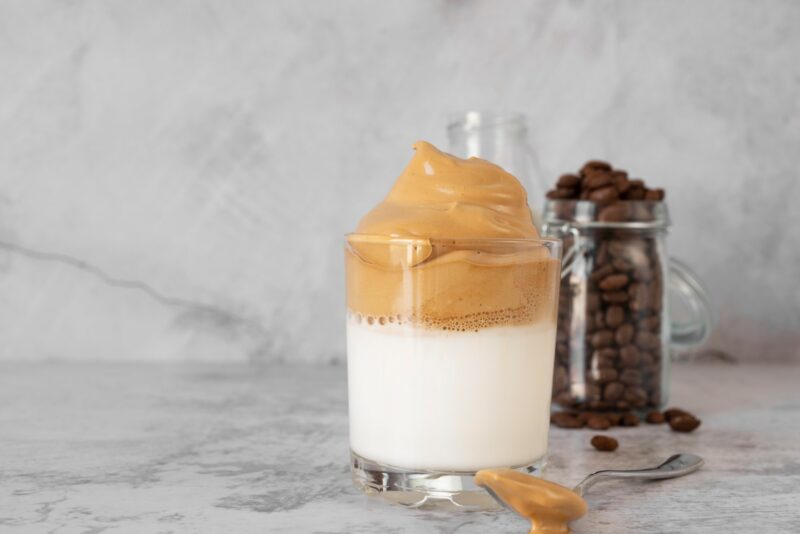 A glass of Dalgona Coffee pictured on a marble top next to a jar of coffee beans.