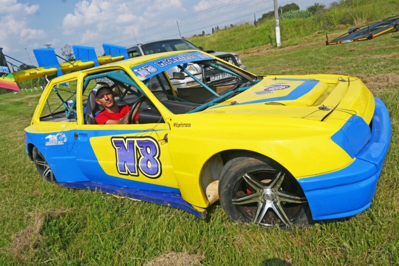 A yellow and blue oval track track car is pictured with its driver inside it.
