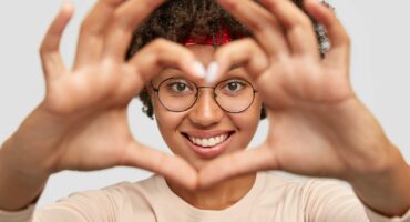 Close up shot of cheerful carefree charming dark skinned girl makes heart shape hand sign over face, demonstrates her love on distance, focus on hands, cares about someone. Romantic affection.