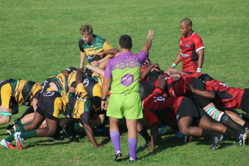 A photo taken of two rival rugby teams during a scrum on a rugby field. A referee is standing in front of the players.