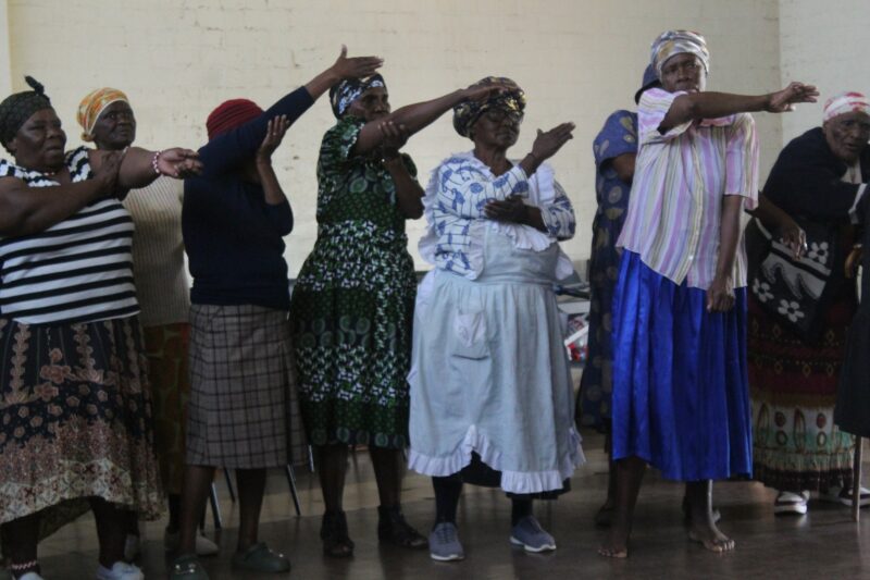 A group of women are photographed dancing inside a community hall.